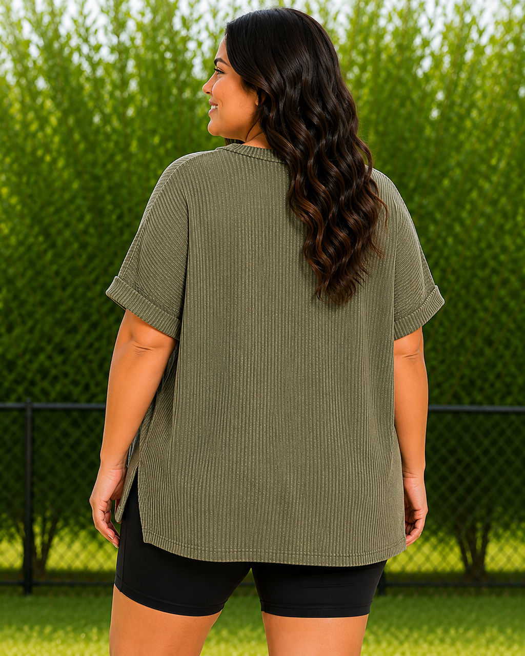 A woman viewed from the back wearing an oversized ribbed short-sleeve top in olive green, paired with black shorts, standing outdoors in front of a green hedge and fence.