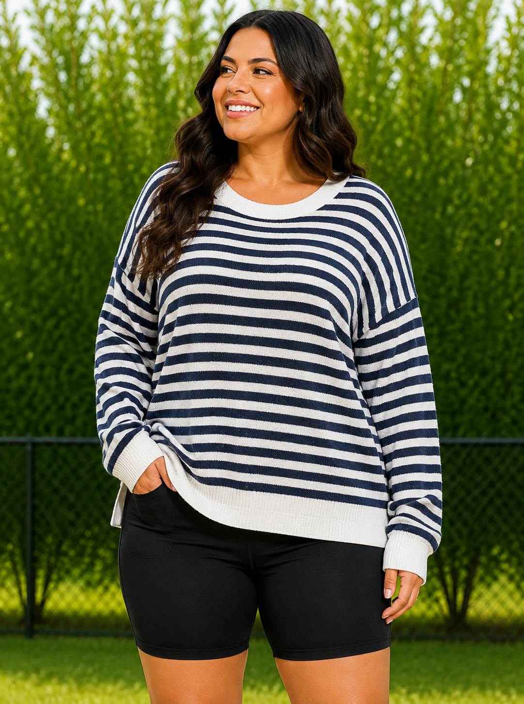 A woman wearing a navy and white horizontally striped long-sleeve knit sweater and black shorts, standing outdoors in front of a green hedge and fence.
