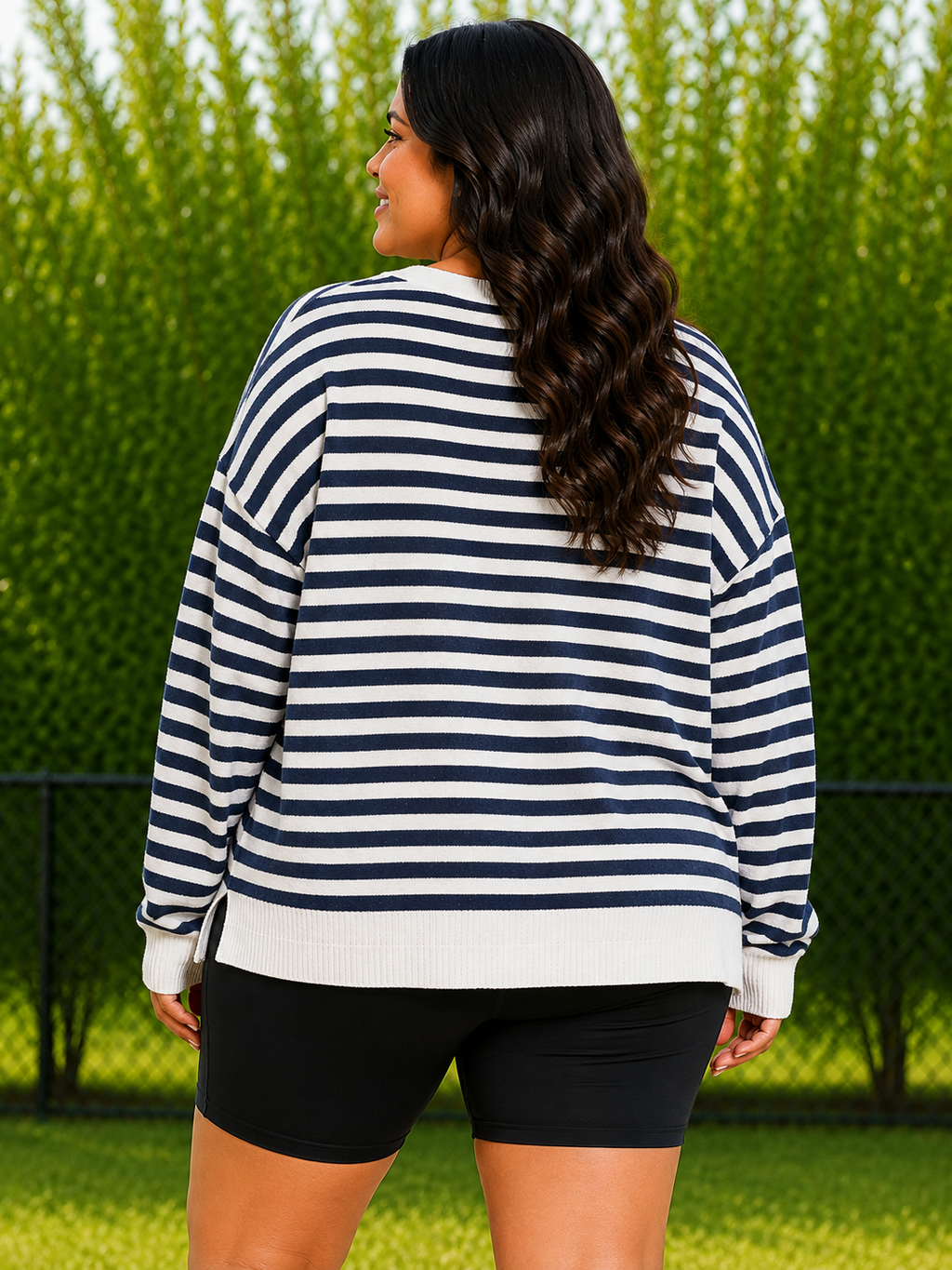 A woman viewed from behind wearing a navy and white horizontally striped long-sleeve sweater and black bike shorts, standing outdoors in front of a green hedge.