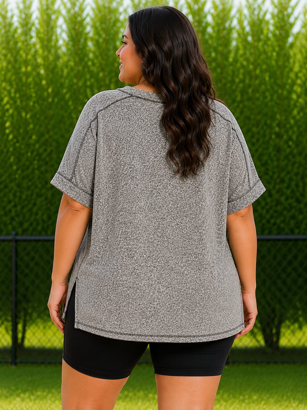 A woman viewed from the back wearing an oversized heather gray short-sleeve top and black bike shorts, standing outdoors in front of a green hedge and fence.