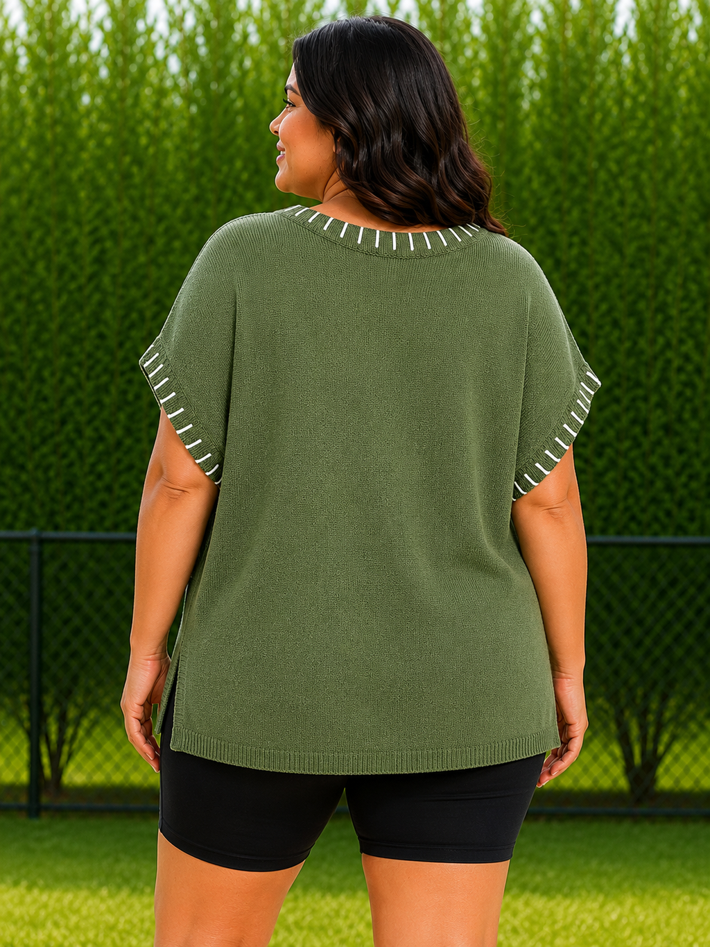 A woman shown from the back wearing an olive green short-sleeve knit top with white contrast stitching along the neckline and sleeve hems, paired with black shorts, set against a green hedge background.