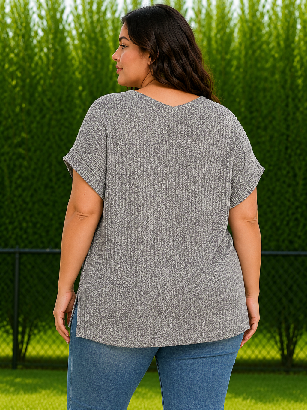 A woman wearing a gray short-sleeve knit top and blue jeans, photographed from the back in an outdoor setting with a green hedge and grass in the background.