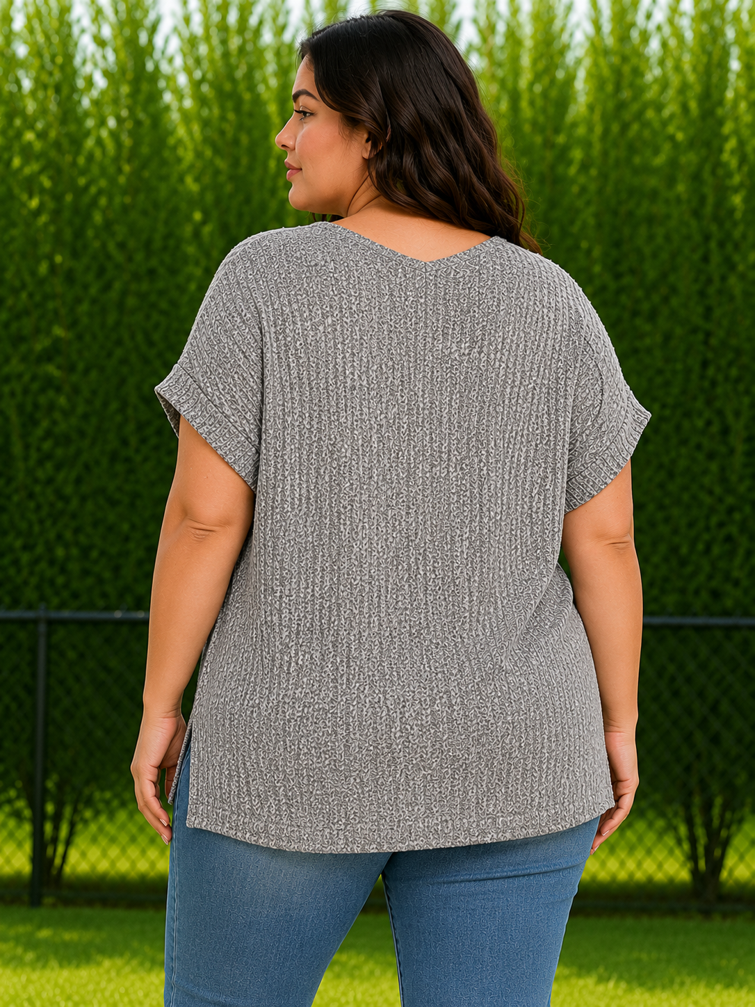A woman wearing a gray short-sleeve knit top and blue jeans, photographed from the back in an outdoor setting with a green hedge and grass in the background.