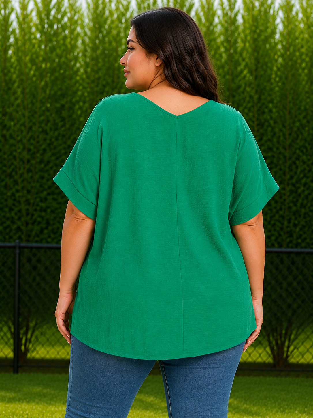 A woman wearing a bright green short-sleeve top and blue jeans, shown from the back in an outdoor setting with a green hedge background.