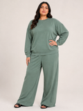 A woman modeling a sage green two-piece loungewear set consisting of a long-sleeve crewneck sweatshirt and matching wide-leg pants, photographed against a neutral light beige studio background.