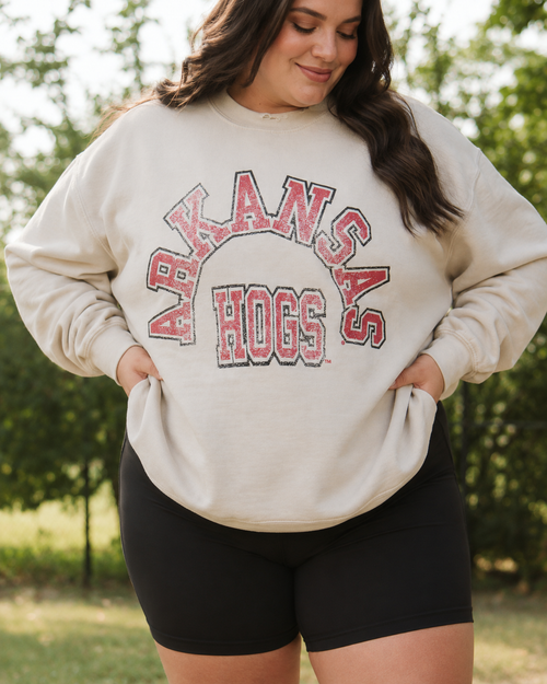 A woman wearing a cream-colored sweatshirt with 'ARKANSAS HOGS' printed in red and black varsity lettering, paired with black bike shorts, standing outdoors in front of a blurred green background.