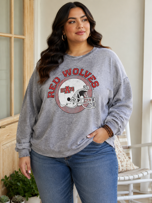 A woman wearing a heather gray long-sleeve sweatshirt with a 'Red Wolves' football helmet graphic on the front, paired with blue jeans, standing in a bright indoor setting.