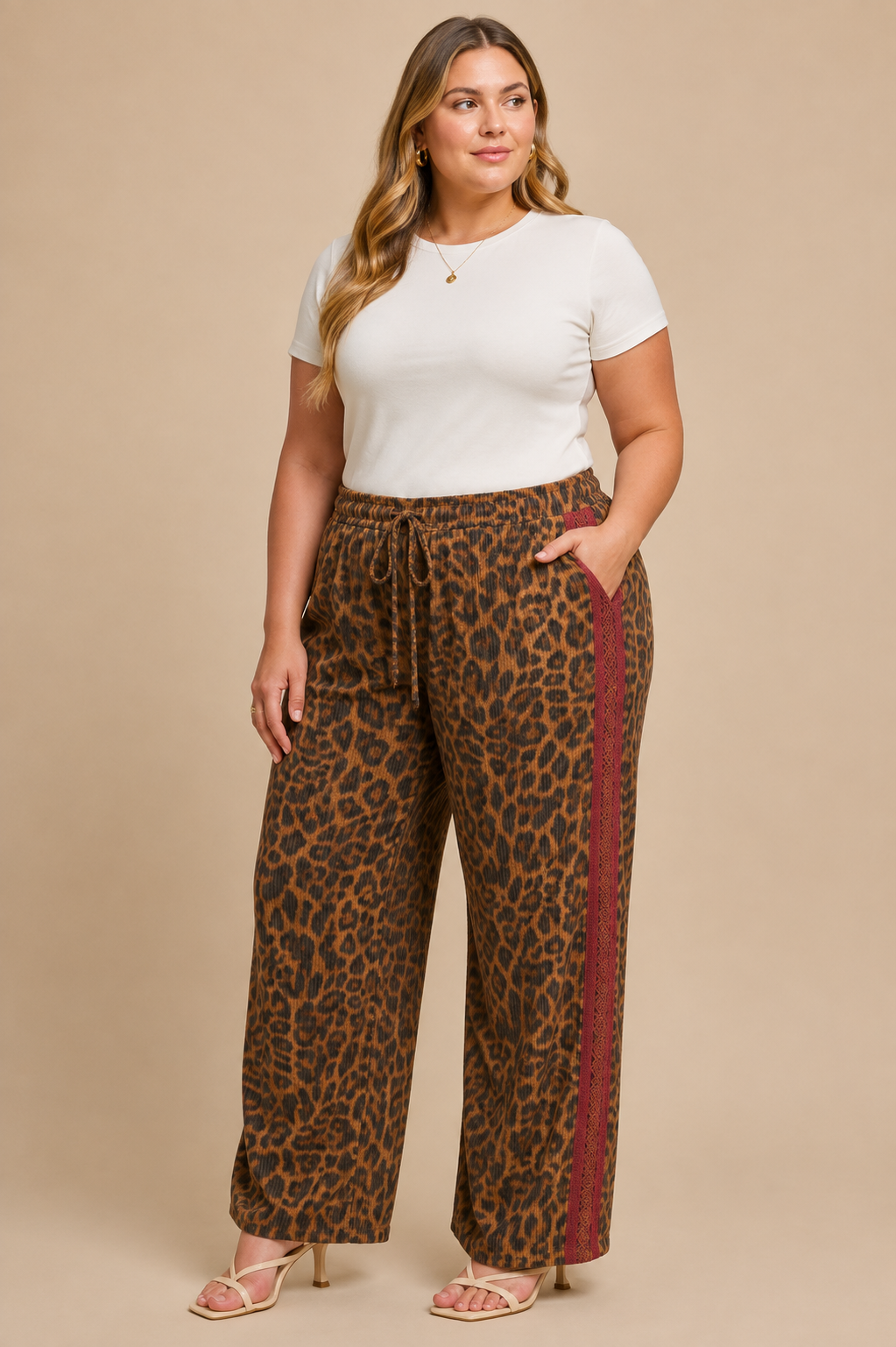 A woman modeling wide-leg pants with a brown and black leopard print and a burgundy side stripe, paired with a white t-shirt and beige heeled sandals, against a solid beige studio background.