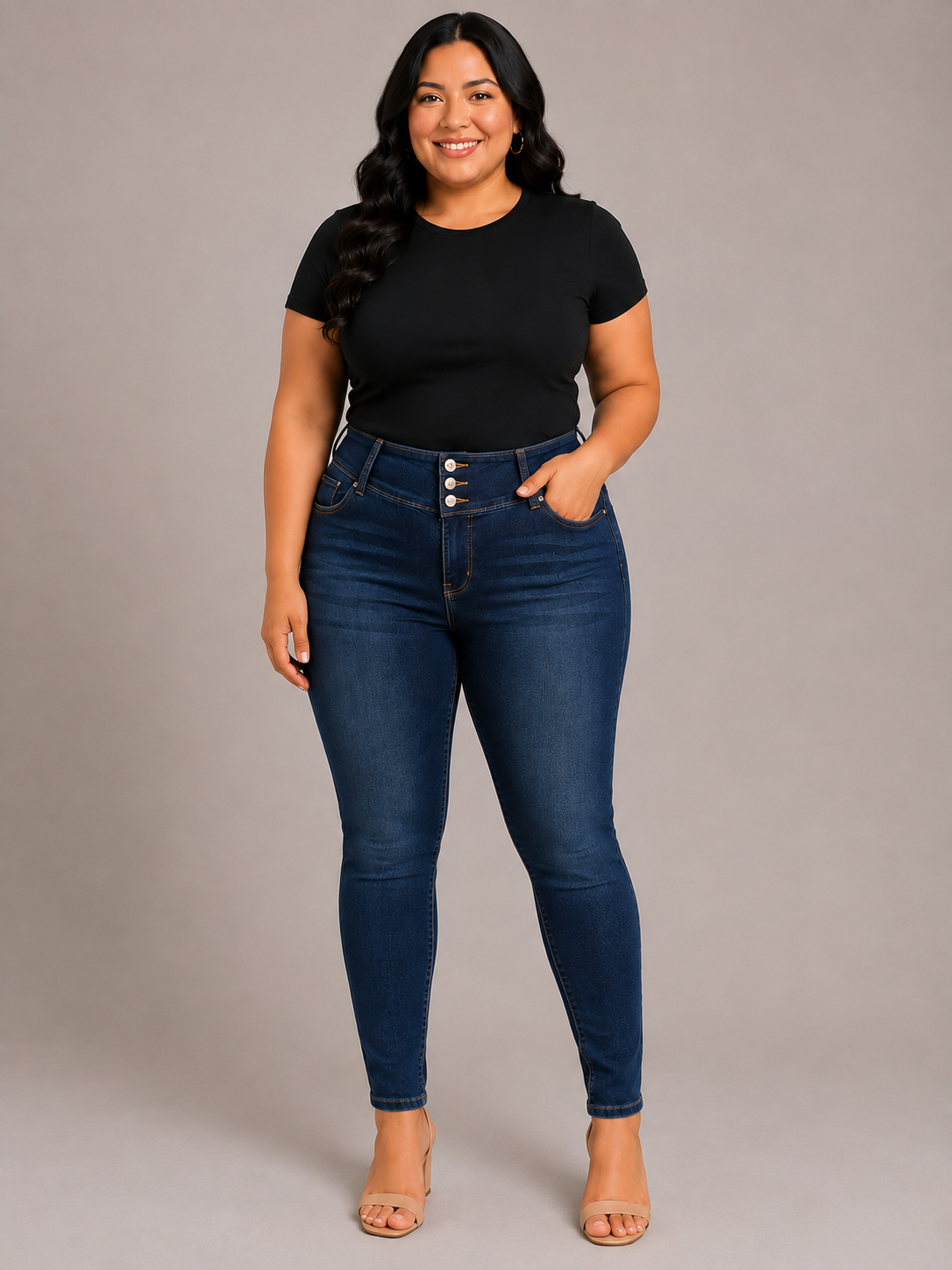 A woman modeling dark blue high-waisted skinny jeans with a button-fly detail, paired with a black t-shirt and beige heeled sandals, against a solid gray studio background.