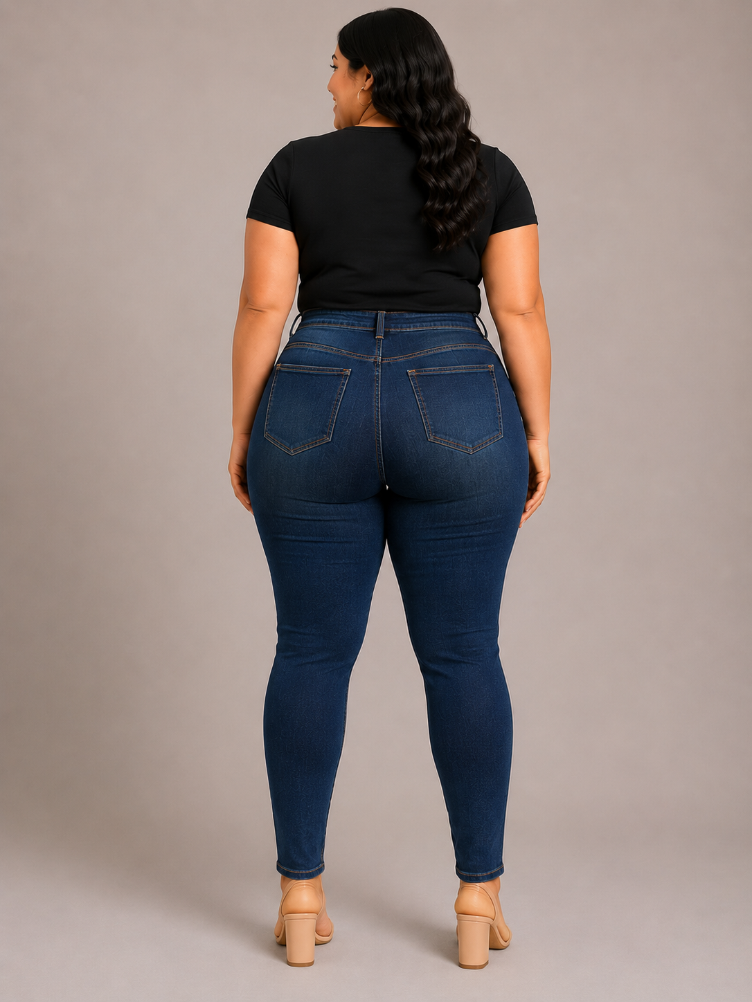 A woman is shown from the back wearing dark blue skinny jeans, a black short-sleeve t-shirt, and beige block-heeled sandals against a solid gray studio background.