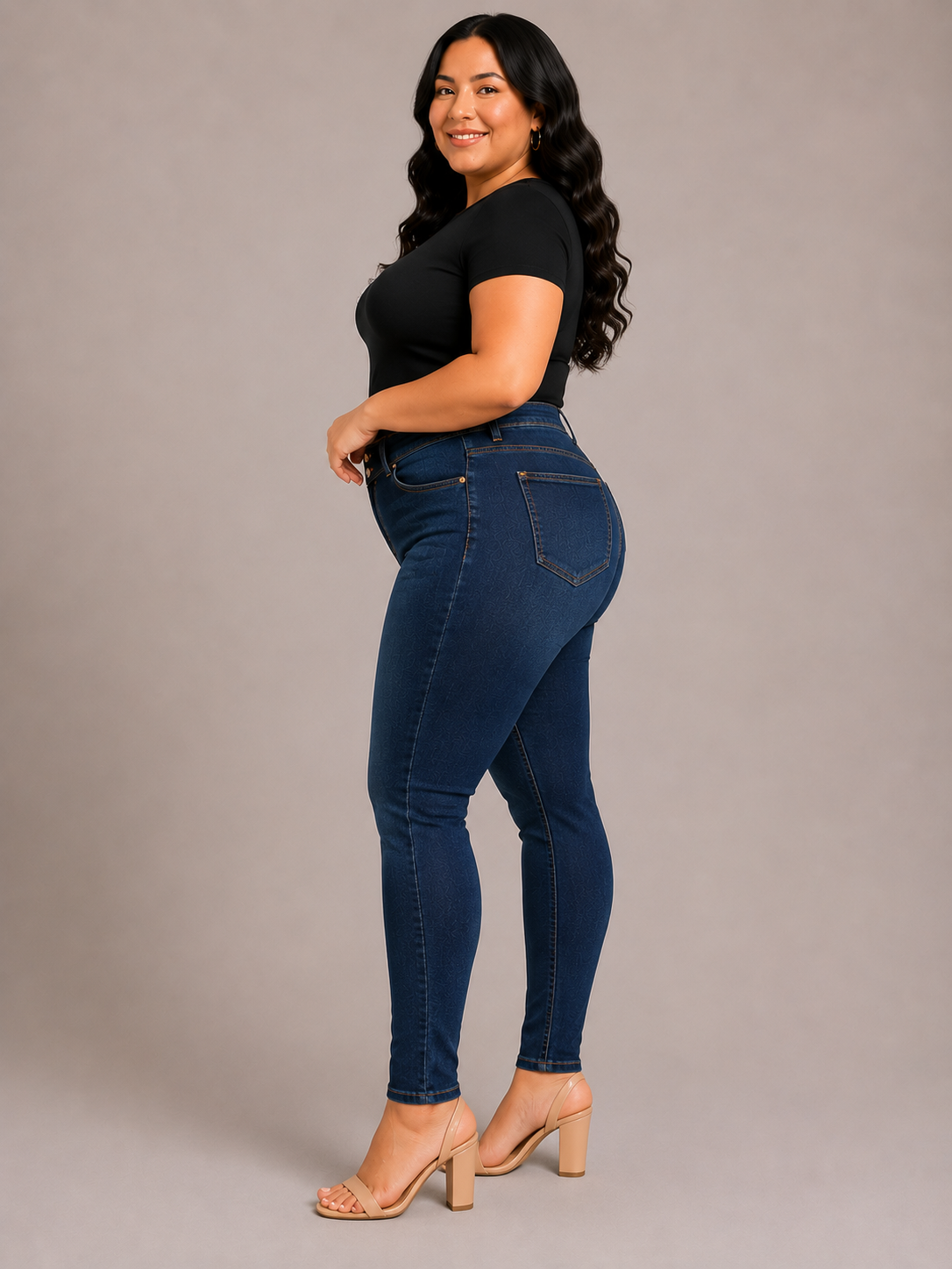 A woman modeling dark blue high-waisted skinny jeans with a black short-sleeve top and beige block-heel sandals, standing against a solid gray studio background.