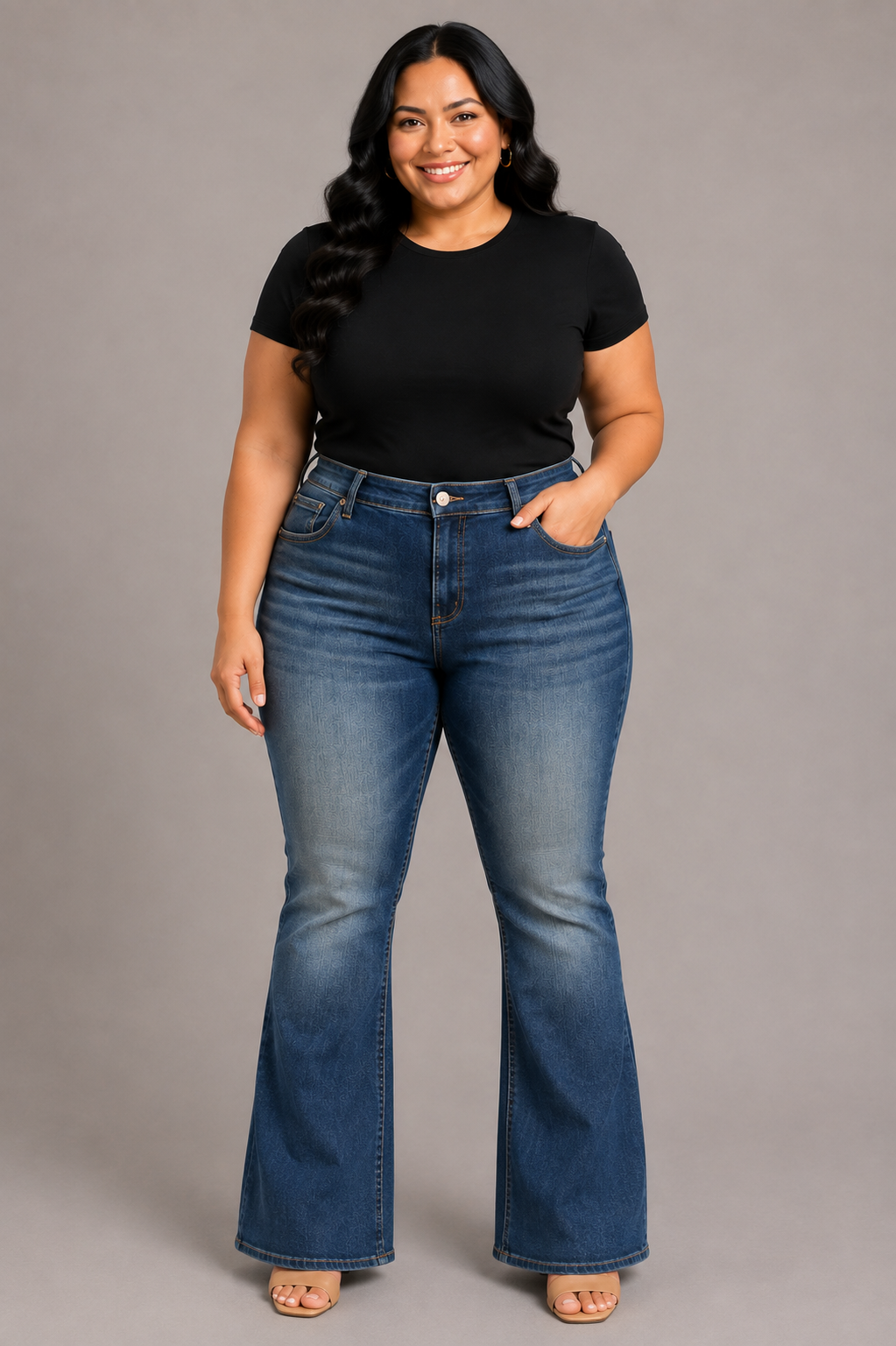 A woman modeling a pair of medium-wash blue flare jeans with a black short-sleeve t-shirt, standing against a solid gray studio background.