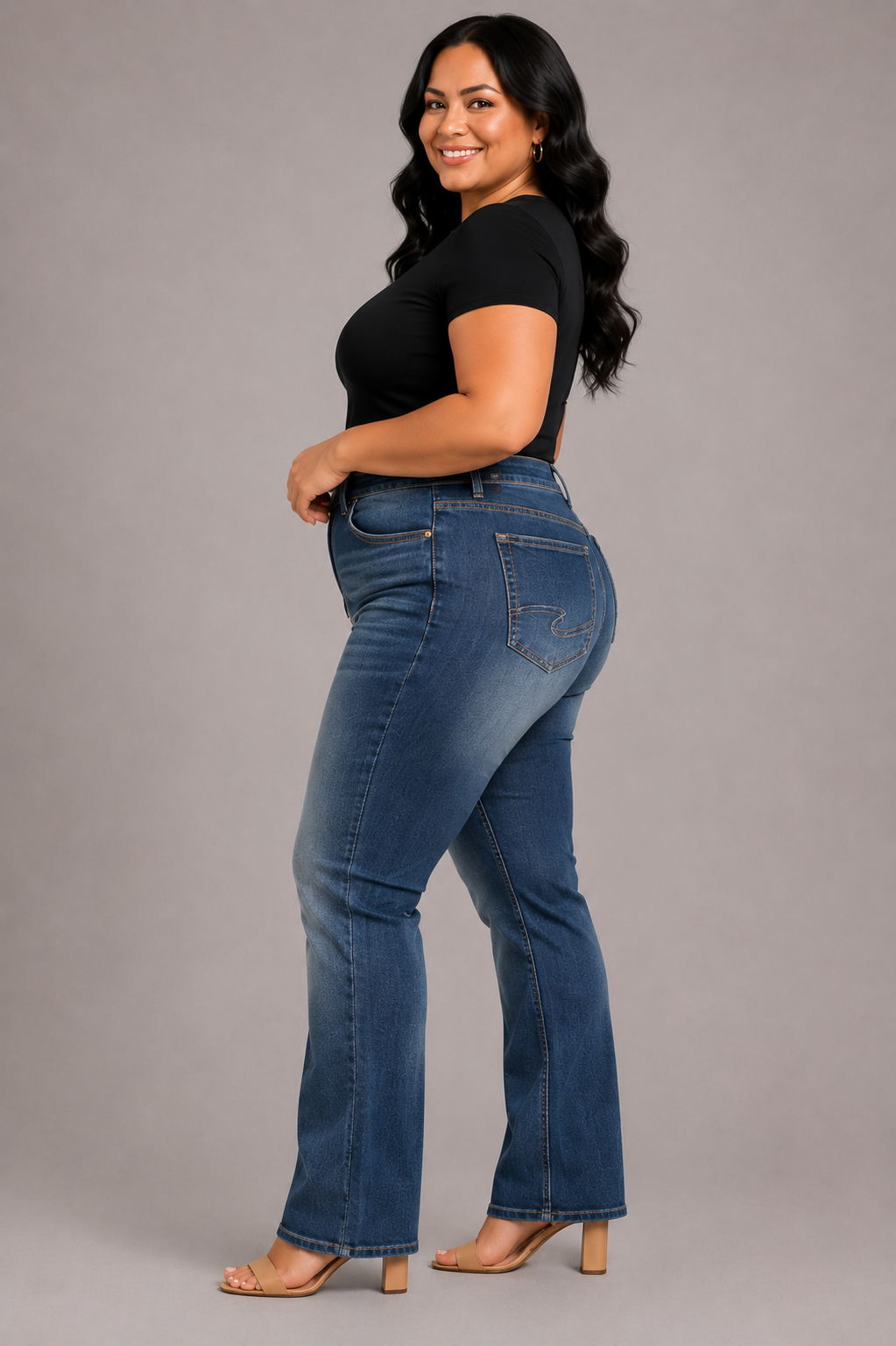 A woman modeling a pair of high-waisted, blue flare jeans with a black short-sleeve top and beige heeled sandals, standing against a solid gray studio background.