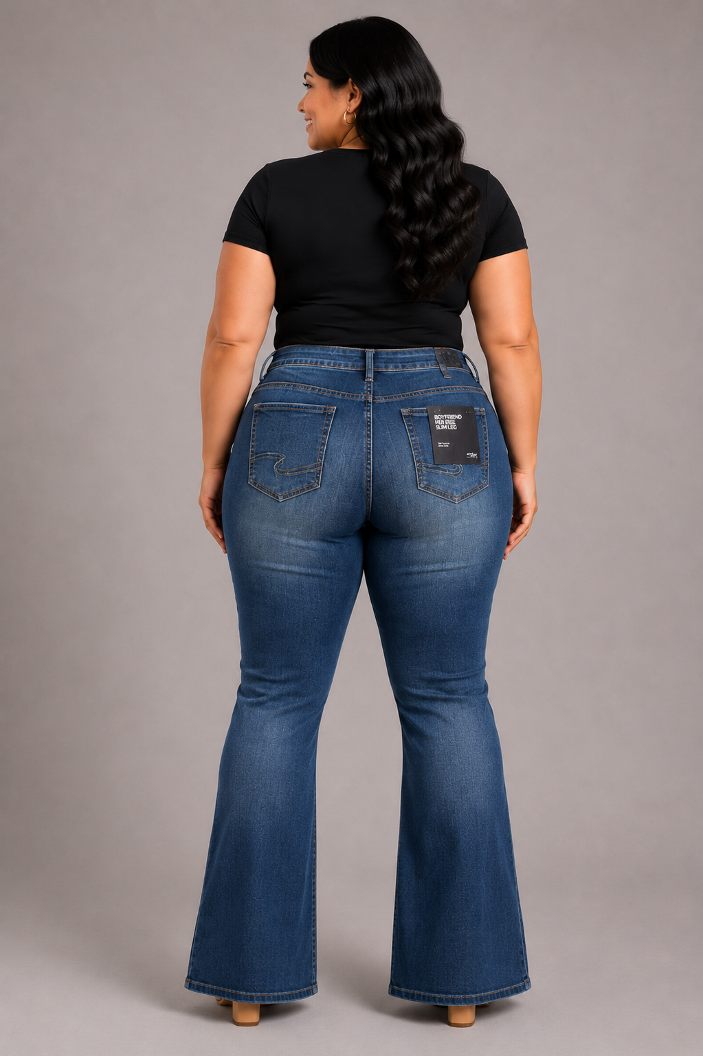 A woman viewed from the back wearing medium-wash blue flare jeans and a black short-sleeve top, standing against a plain gray studio background.