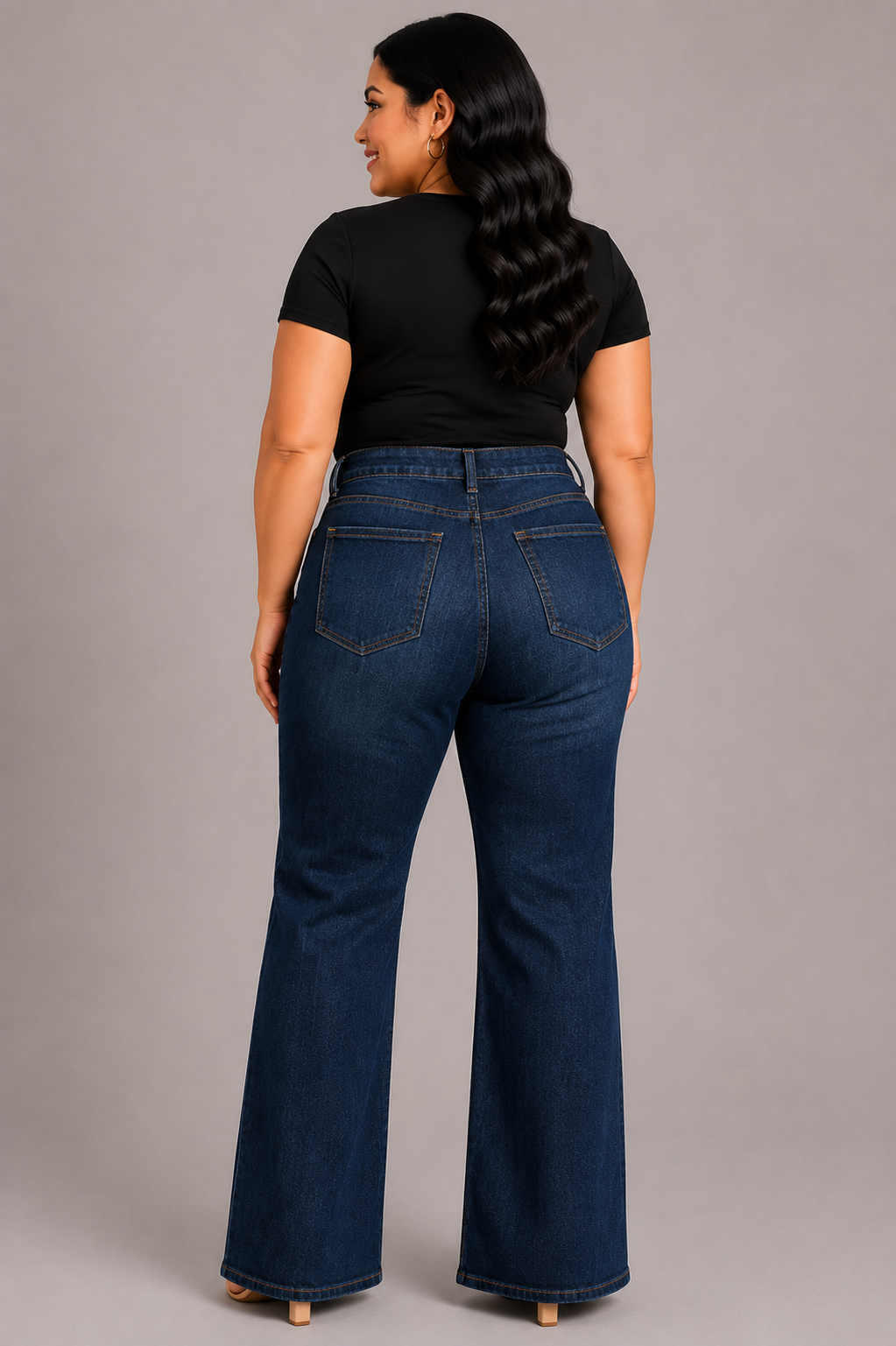 A woman shown from the back wearing dark blue flare jeans and a black short-sleeve top, standing against a solid gray studio background.