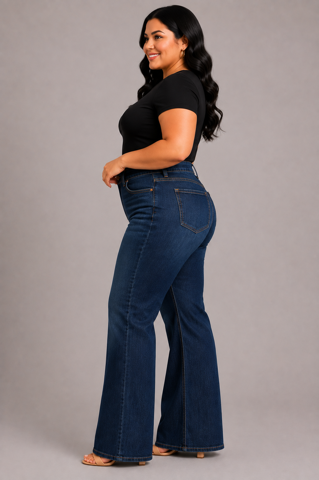 A woman modeling dark blue high-waisted flare jeans with a black short-sleeve top, standing against a solid gray studio background.