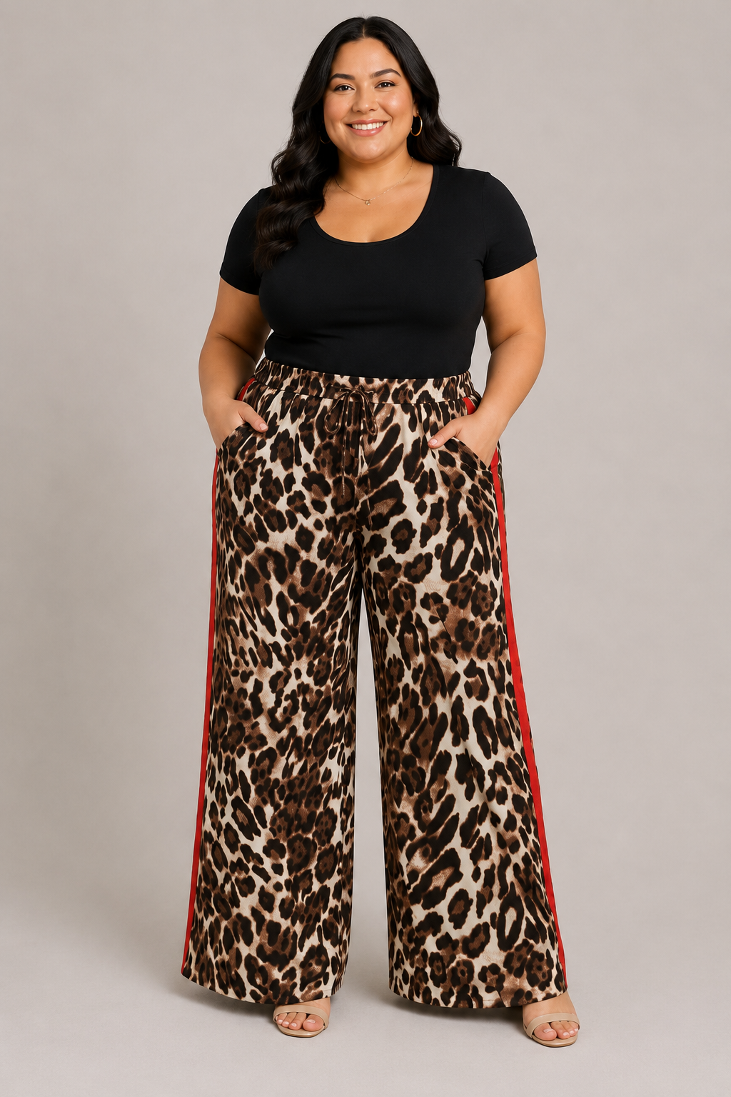 A woman modeling wide-leg leopard print pants with red side stripes, paired with a black short-sleeve top, against a solid light gray studio background.