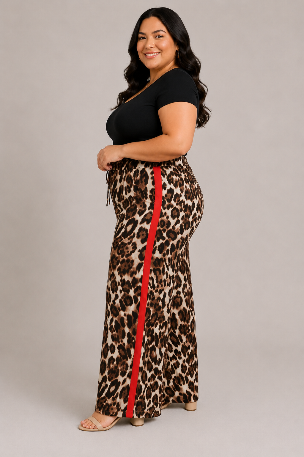 A woman modeling leopard-print maxi pants with a bold red side stripe, paired with a black short-sleeve top, against a solid light gray studio background.