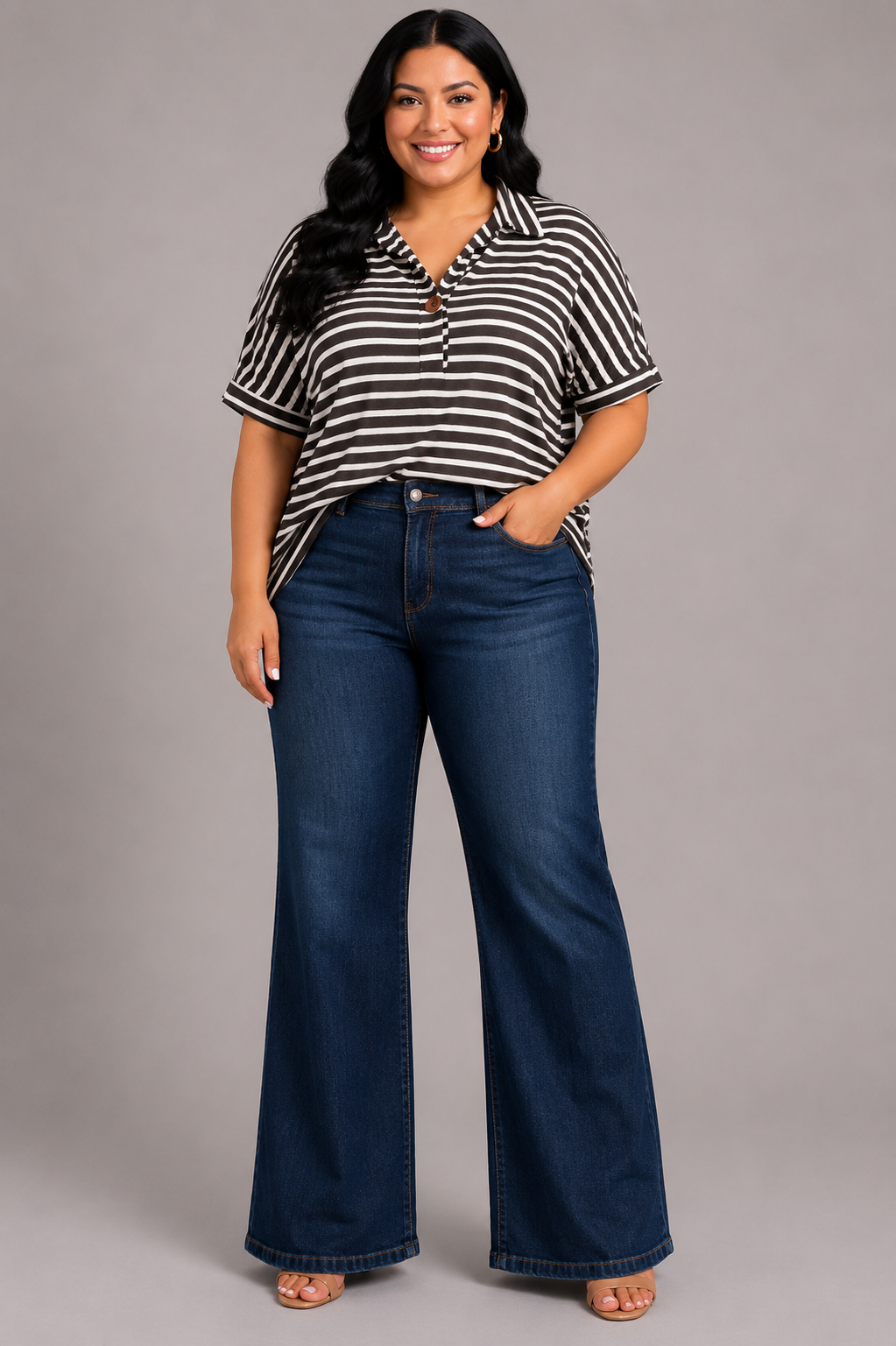 A woman modeling a black and white horizontally striped short-sleeve polo shirt with dark blue flare jeans, standing against a solid gray studio background.
