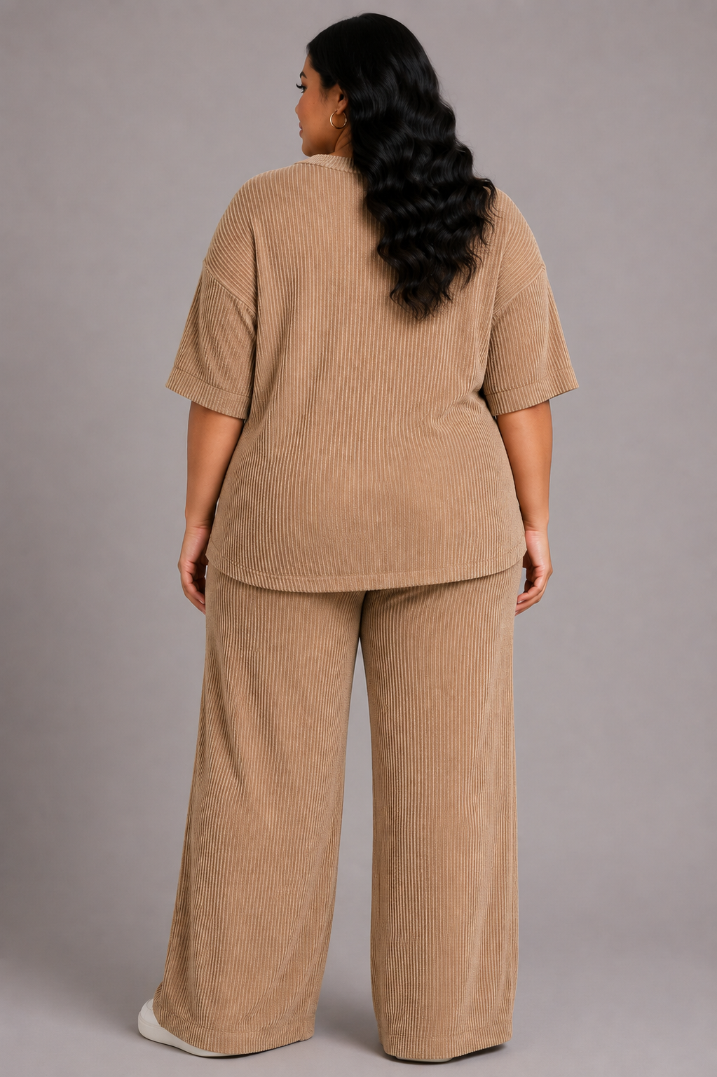 A woman stands with her back to the camera wearing a matching beige corduroy lounge set consisting of a short-sleeve top and wide-leg pants, photographed against a solid gray studio background.