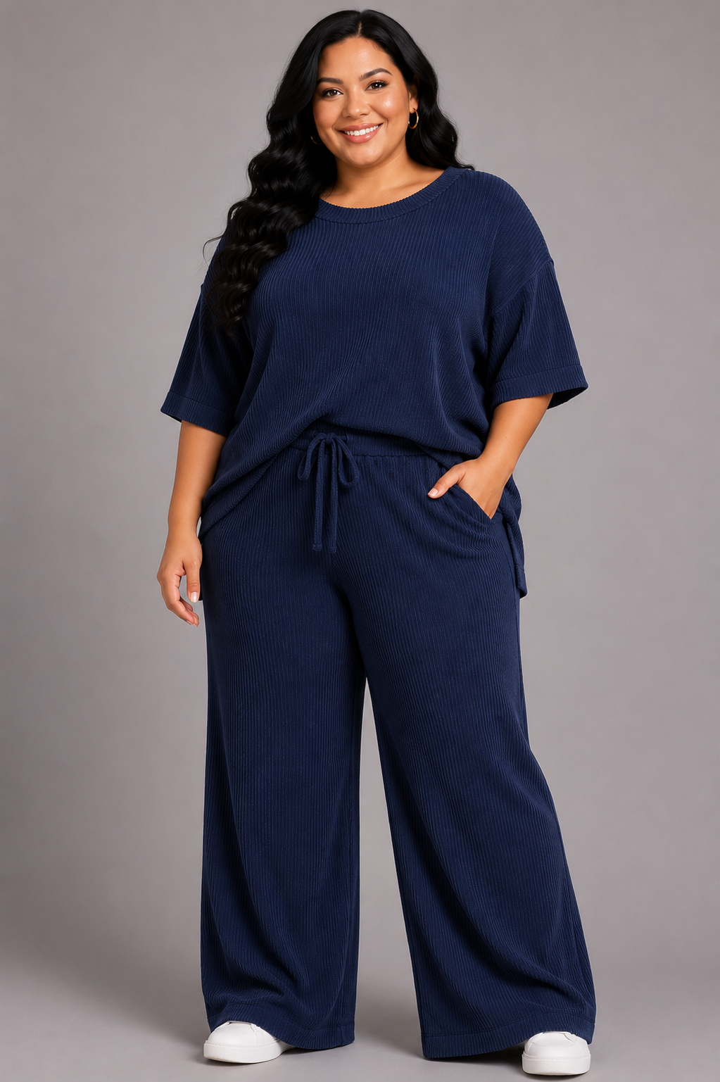 A woman modeling a matching navy blue lounge set consisting of a short-sleeve top and wide-leg pants, photographed against a solid gray studio background.