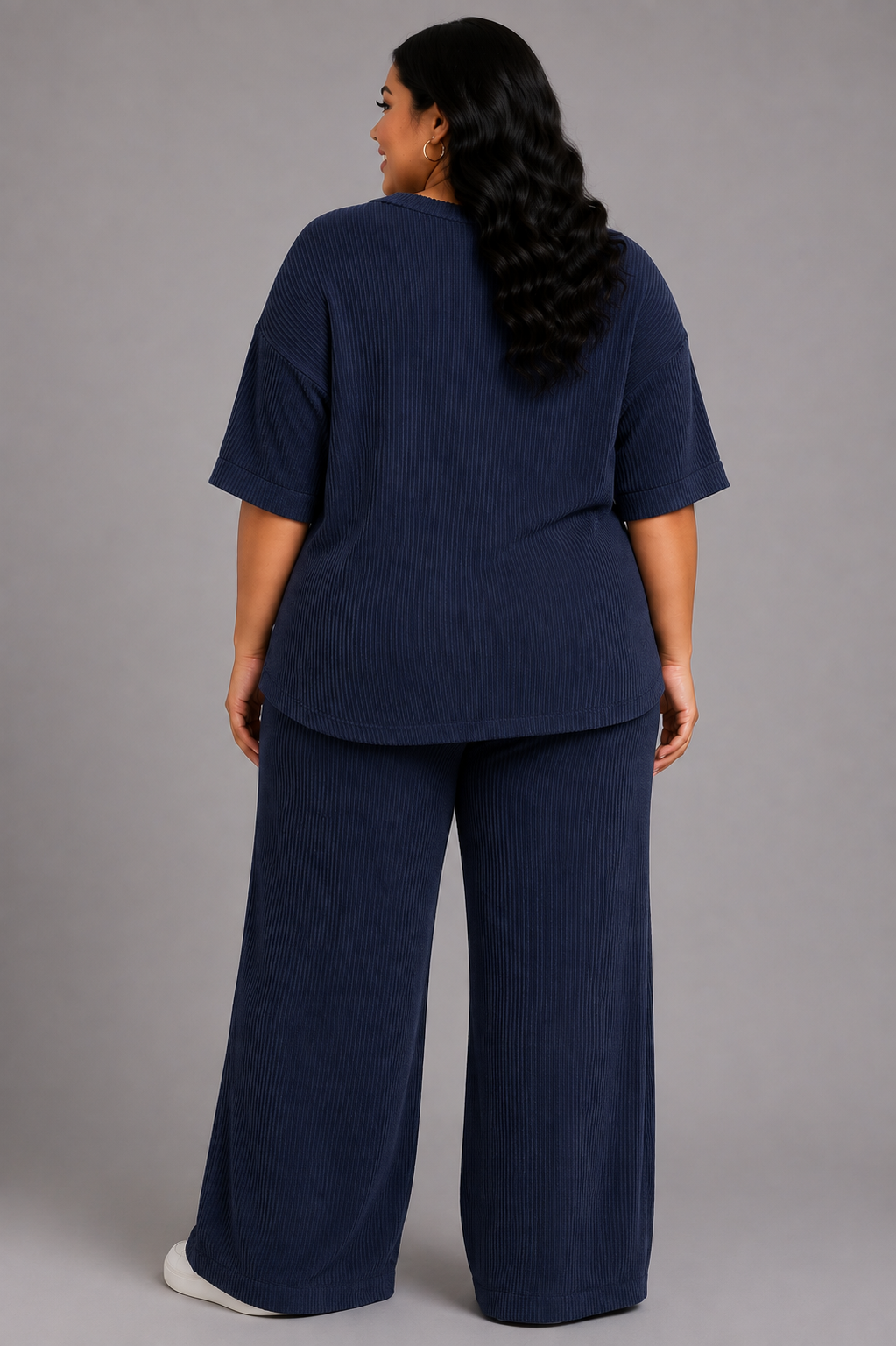 A woman wearing a matching navy blue ribbed lounge set, consisting of a short-sleeve top and wide-leg pants, shown from the back against a solid gray studio background.