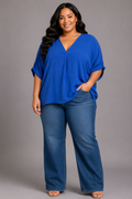 A woman modeling a bright blue short-sleeve V-neck top with wide legs, paired with dark blue jeans, standing against a solid gray studio background.