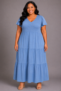 A woman modeling a light blue tiered maxi dress with flutter sleeves and a V-neckline, standing against a solid gray studio background.