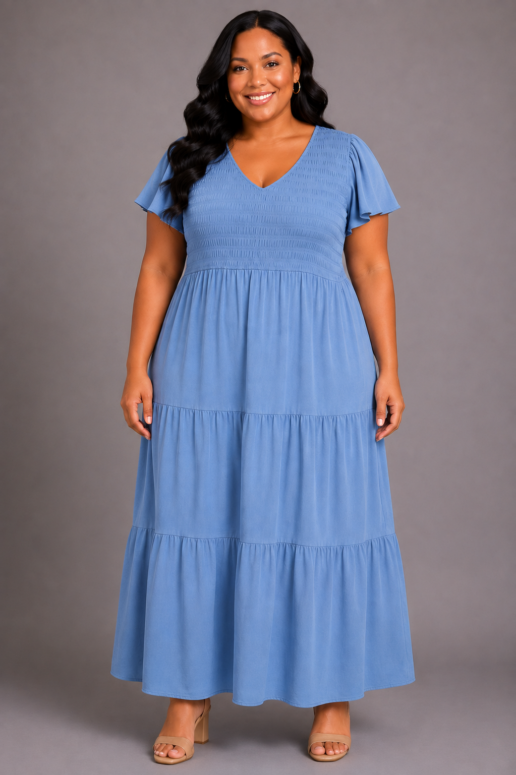 A woman modeling a light blue tiered maxi dress with flutter sleeves and a V-neckline, standing against a solid gray studio background.