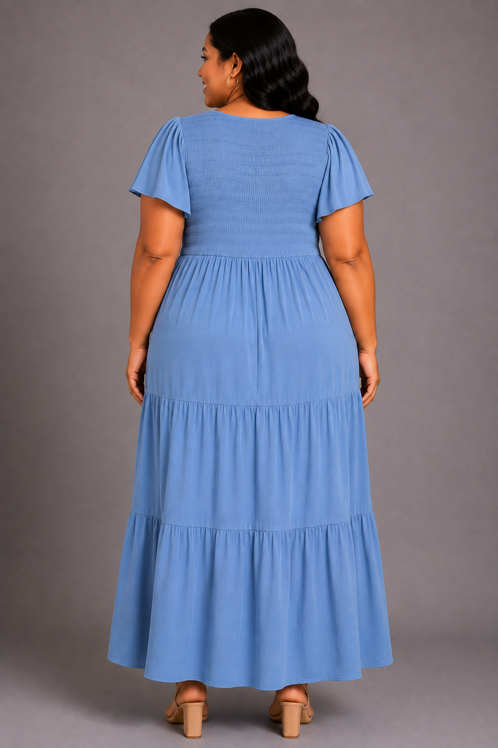 A woman viewed from the back wearing a light blue tiered maxi dress with short puff sleeves, standing against a plain gray studio background.