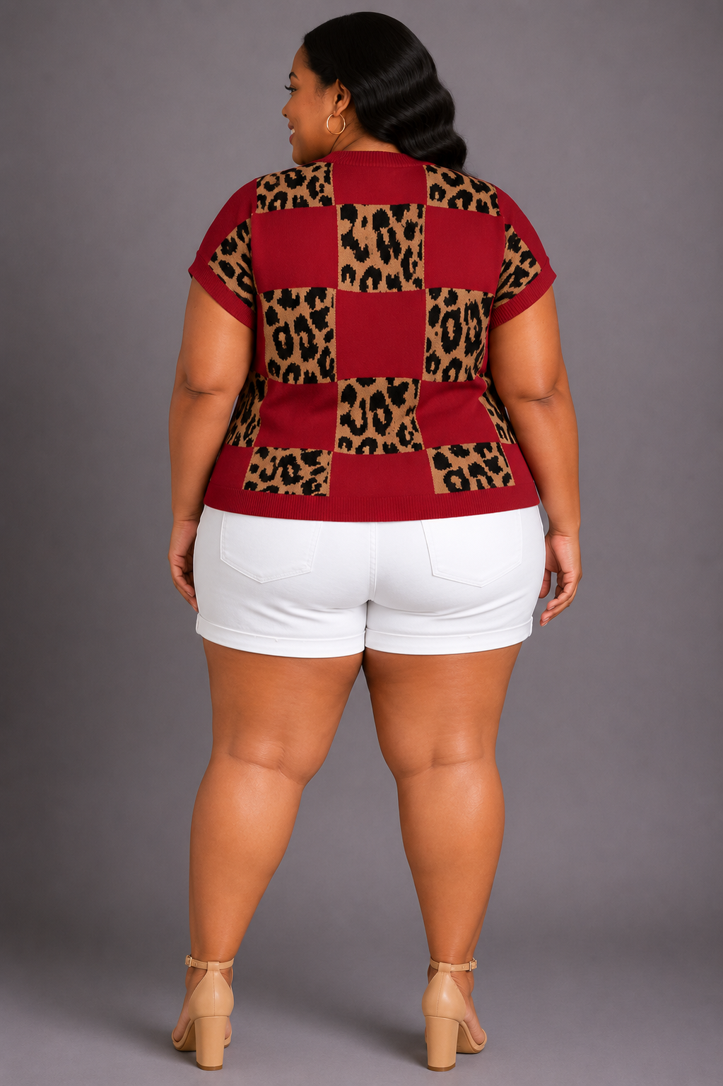 A woman shown from the back wearing a short-sleeve top with a red and leopard print checkerboard pattern, white shorts, and beige heels against a solid gray studio background.
