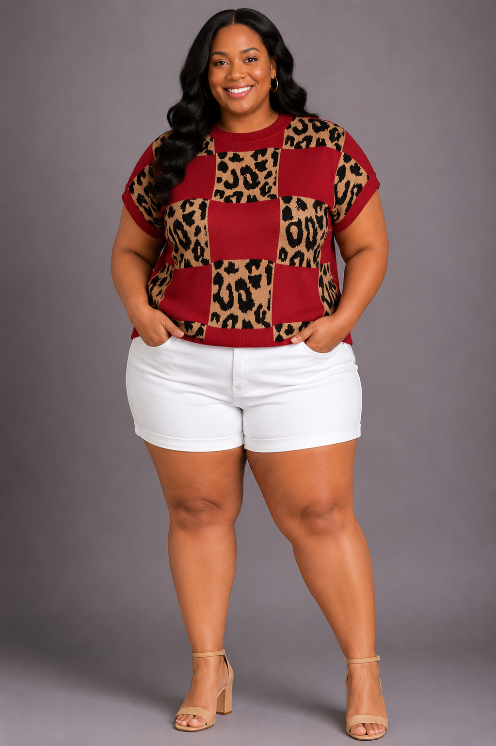 A woman modeling a short-sleeve knit top with a checkered pattern of red squares and leopard print squares, paired with white shorts and beige high-heeled sandals, against a solid gray studio background.
