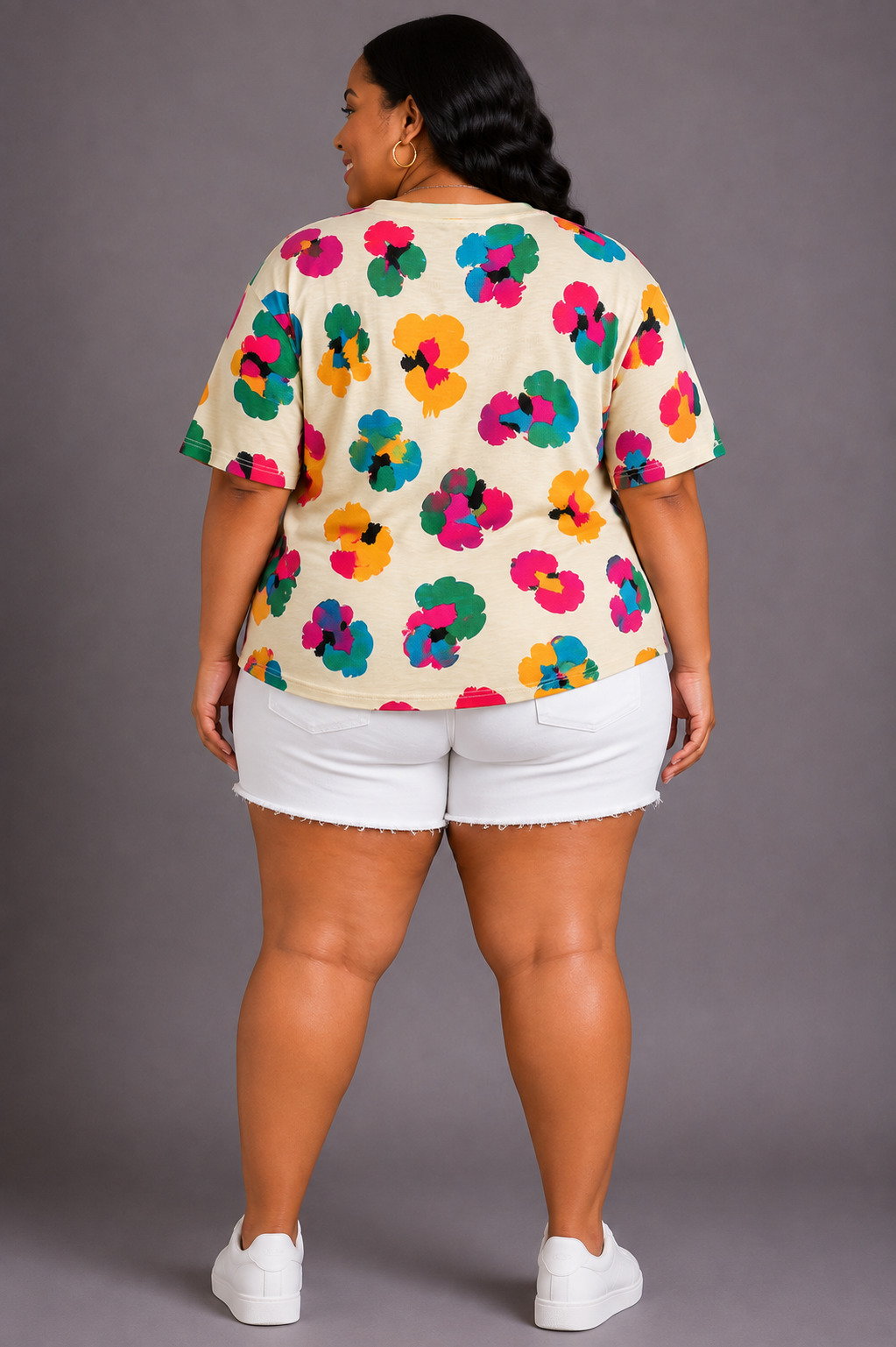 A plus-size woman is shown from the back wearing a cream-colored short-sleeve t-shirt with a vibrant multicolor floral pattern, paired with white denim shorts and white sneakers, against a solid gray studio background.