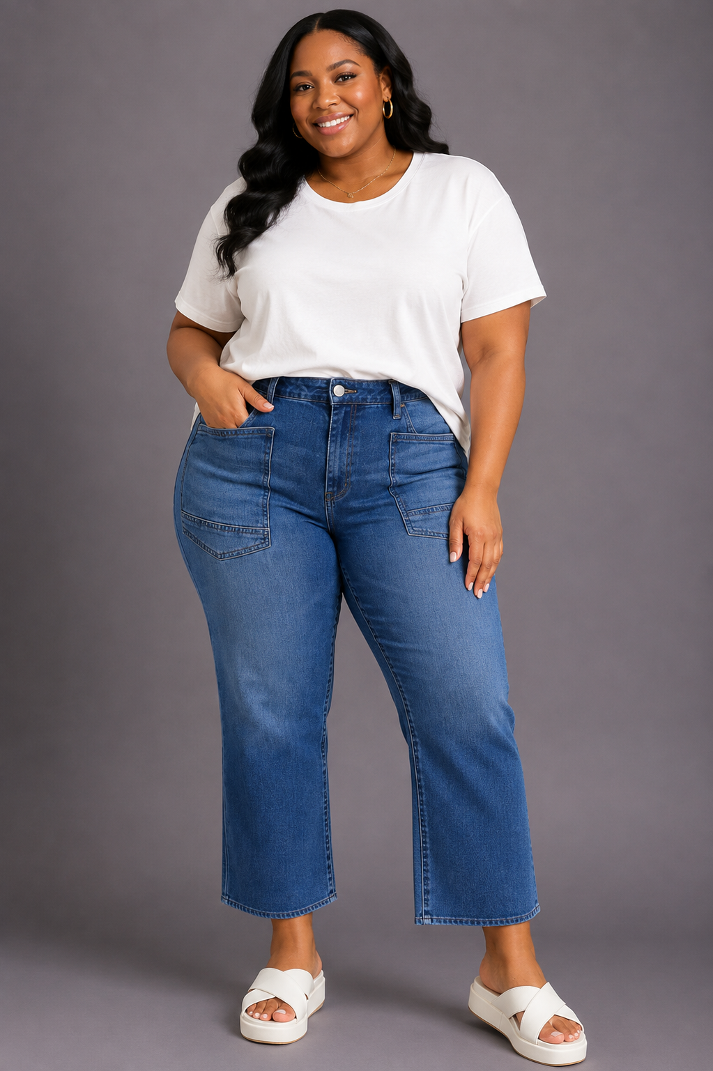 A woman modeling a pair of blue cropped flare jeans with a white t-shirt and white slide sandals, standing against a solid gray studio background.