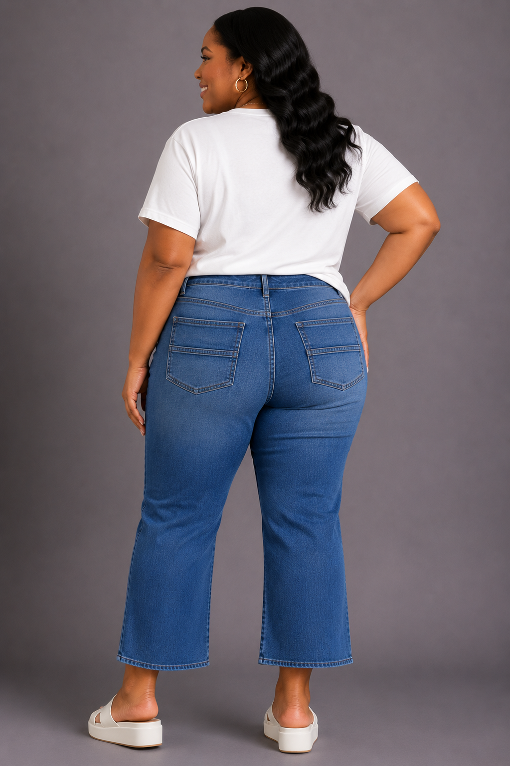 A woman shown from the back wearing blue cropped flare jeans, a white t-shirt, and white slide sandals against a solid gray studio background.