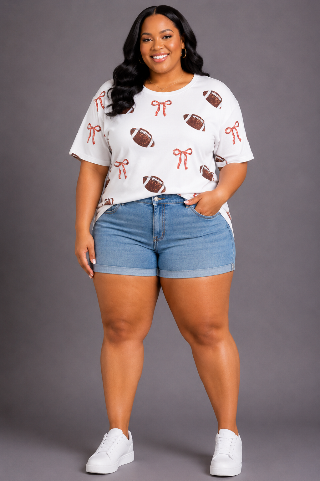 A woman modeling a white short-sleeve t-shirt with a pattern of brown footballs and red bows, paired with blue denim shorts and white sneakers, against a solid gray studio background.