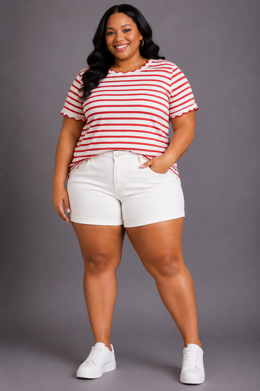 A woman modeling a red and white striped short-sleeve t-shirt with scalloped edges, paired with white denim shorts and white sneakers, against a solid gray studio background.