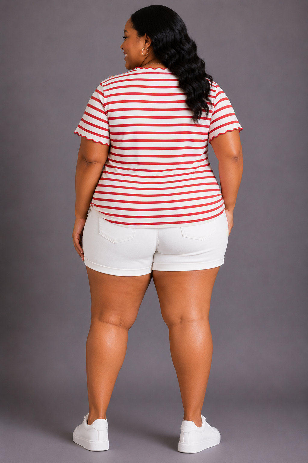 A woman shown from the back wearing a red and white horizontally striped short-sleeve top with scalloped edges, white shorts, and white sneakers, standing against a solid gray studio background.