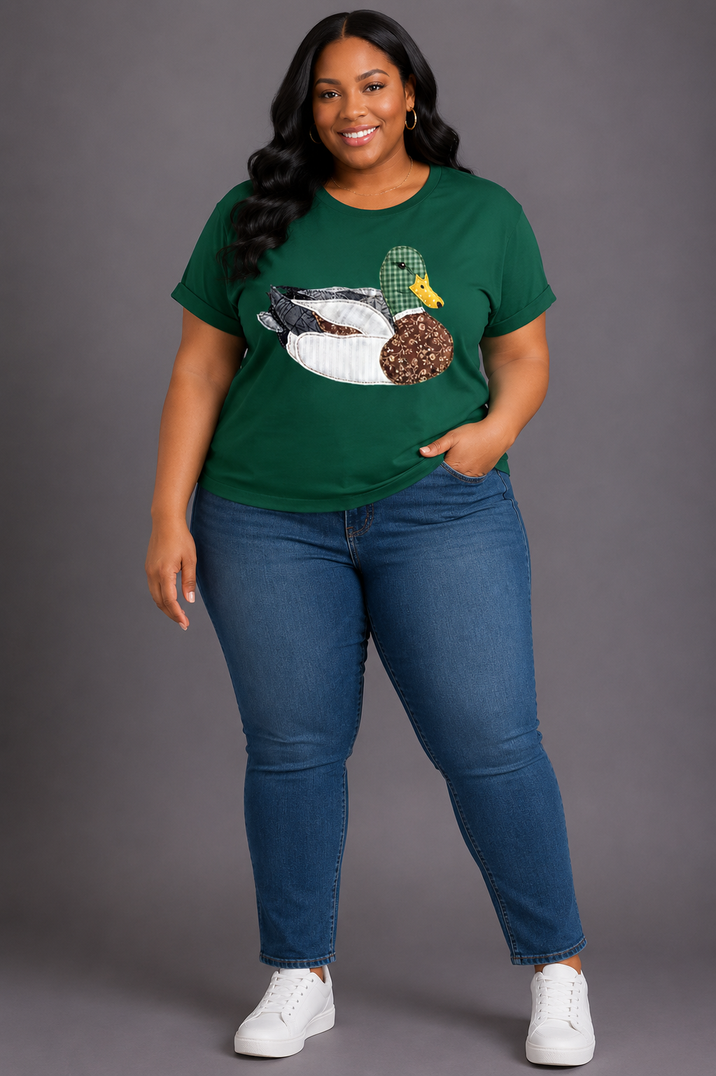 A woman modeling a dark green graphic t-shirt with a duck and sausage design, paired with blue jeans and white sneakers, against a solid gray studio background.
