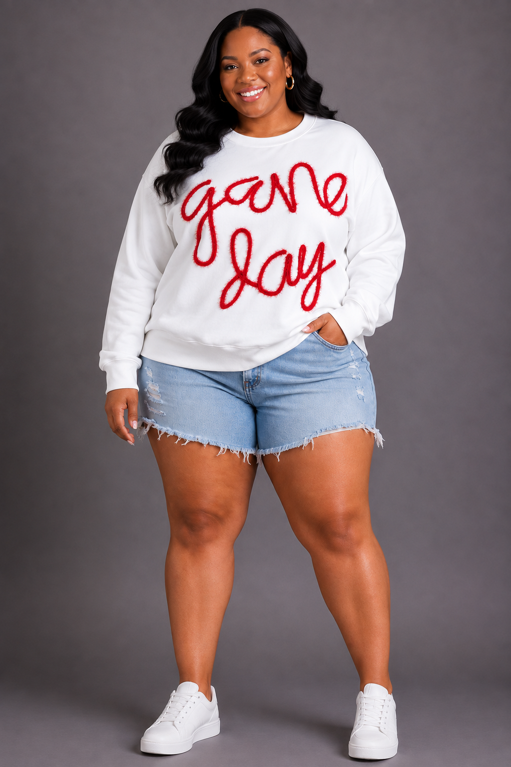 A woman modeling a white crewneck sweatshirt with 'game day' written in red cursive across the front, paired with light blue denim shorts and white sneakers, against a solid gray studio background.