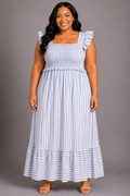 A woman modeling a sleeveless maxi dress with ruffled shoulder straps, a smocked bodice, and vertical light blue and white stripes, standing against a solid gray studio background.