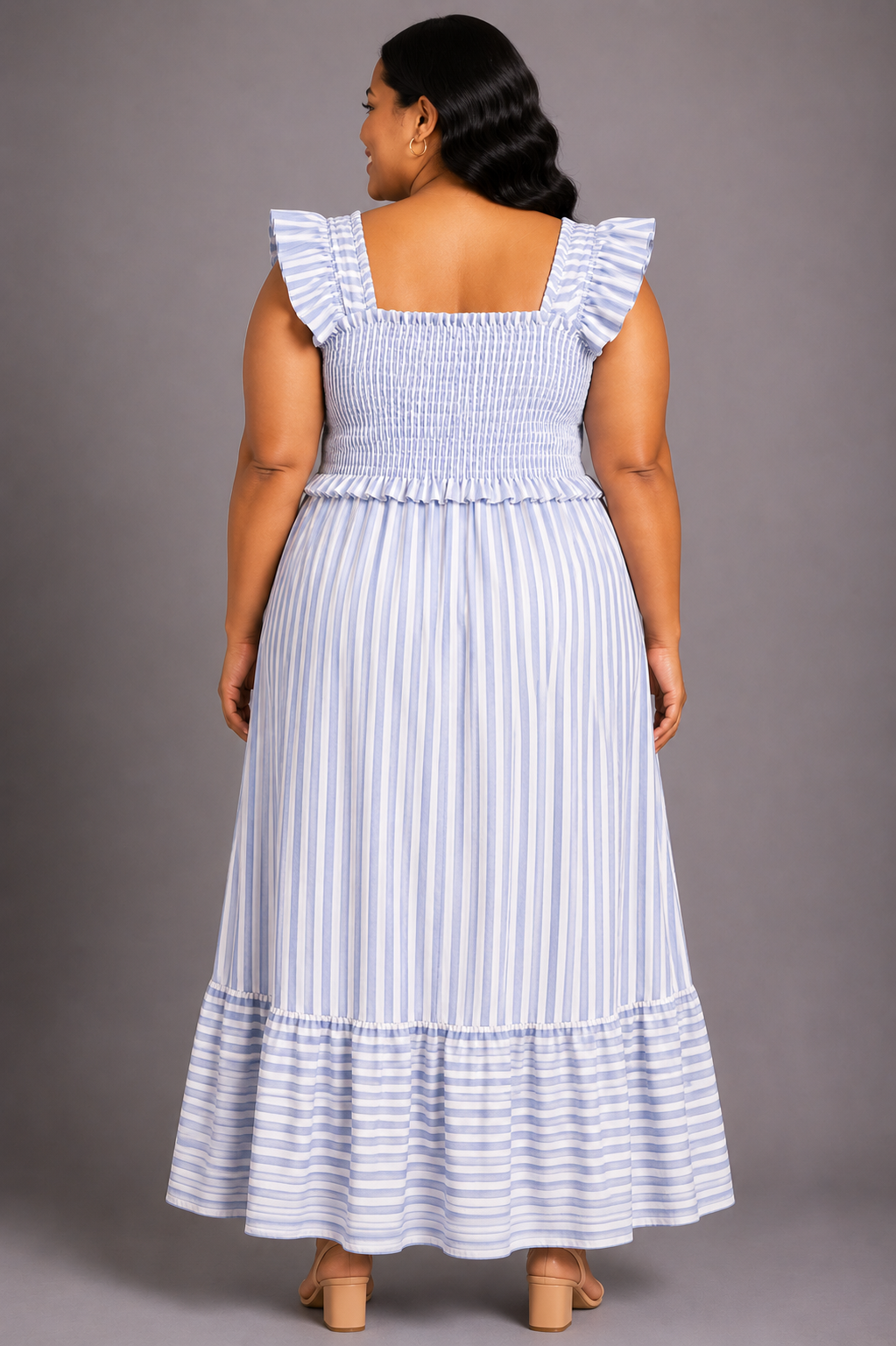 A woman shown from the back wearing a sleeveless maxi dress with ruffled shoulder straps, a smocked bodice, and vertical light blue and white stripes, photographed against a plain gray studio background.