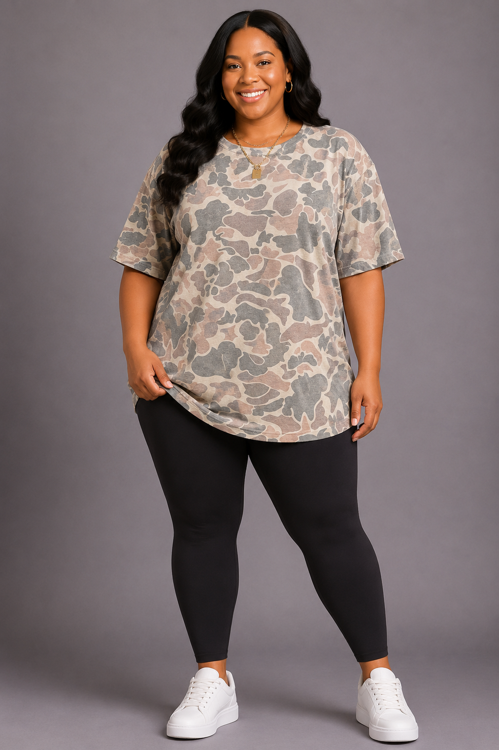 A woman modeling a short-sleeve crewneck t-shirt with a beige, brown, and gray camouflage pattern, paired with black leggings and white sneakers, against a solid gray studio background.