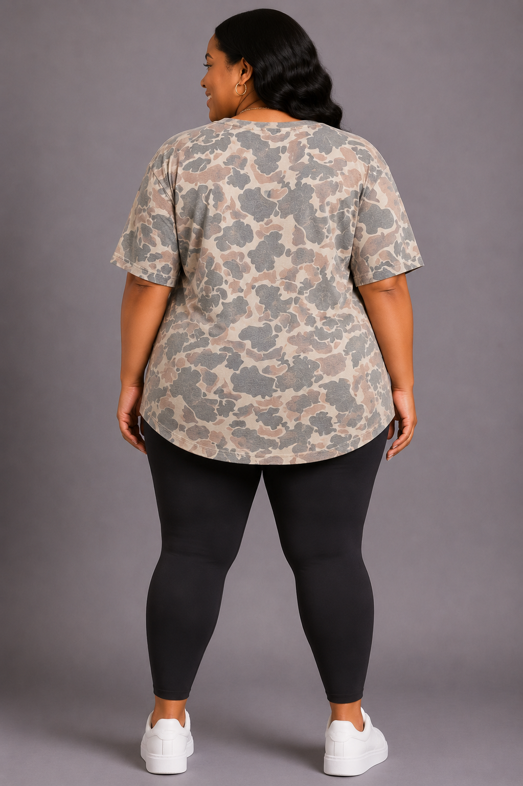 A woman shown from the back wearing a short-sleeve t-shirt with a beige and gray abstract camouflage pattern, paired with black leggings and white sneakers, against a solid gray studio background.
