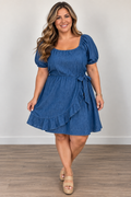 A woman modeling a blue short-sleeve dress with puff sleeves, a square neckline, a tie waist, and a ruffled hem, standing against a plain light gray wall on a wood floor.