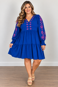 A woman modeling a royal blue tiered dress with pink floral embroidery on the neckline, sleeves, and cuffs, paired with beige platform sandals against a neutral studio background.