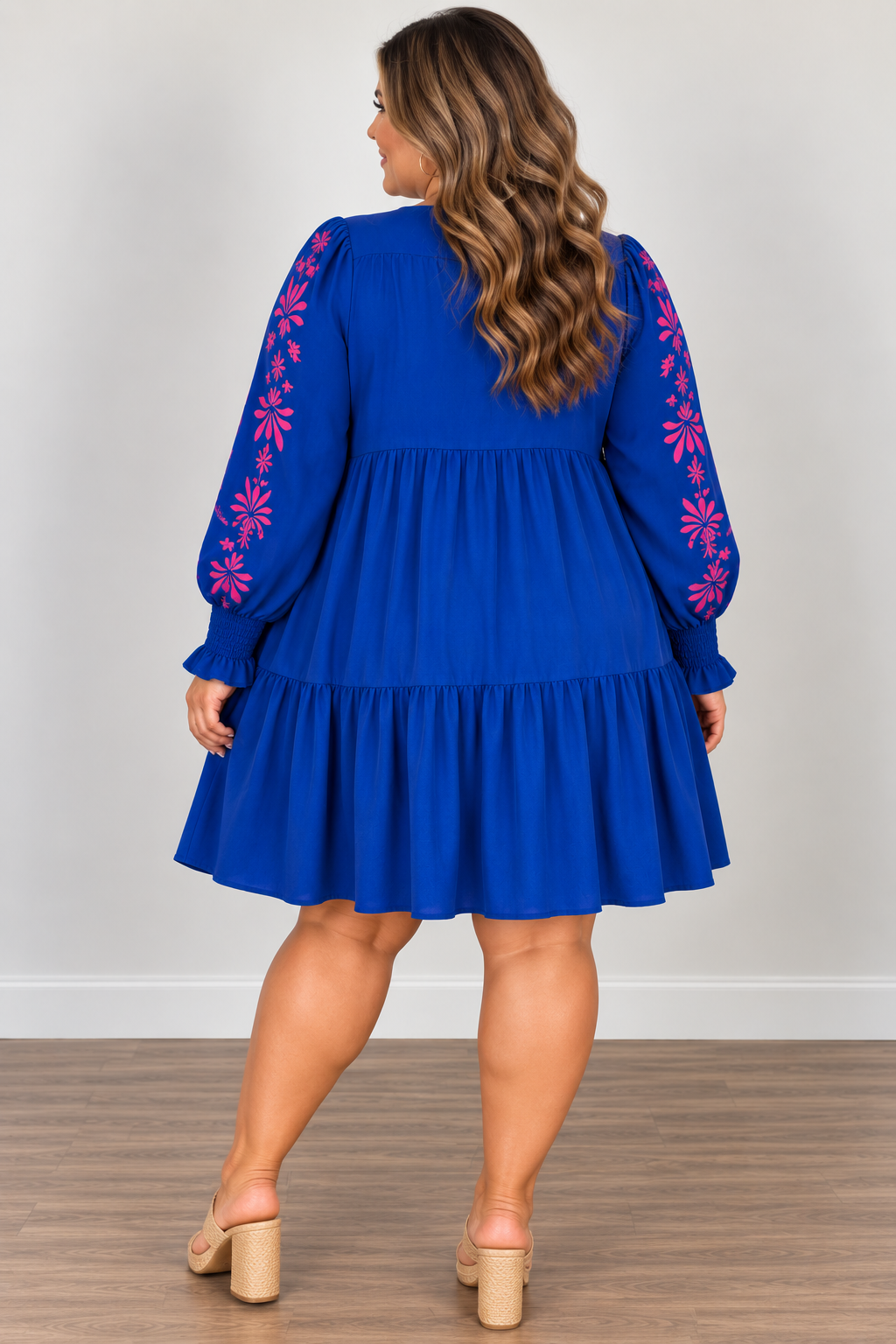 A woman viewed from the back wearing a bright royal blue tiered dress with long sleeves featuring pink floral embroidery, paired with beige block-heel mules, set against a neutral indoor background.