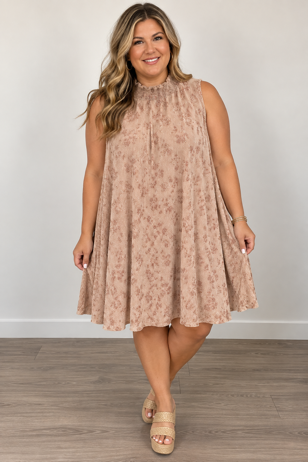 A woman modeling a sleeveless, high-neck, knee-length pleated dress in a dusty rose-beige color with a subtle tonal floral pattern, paired with beige platform sandals, set against a plain light gray wall and wood floor.