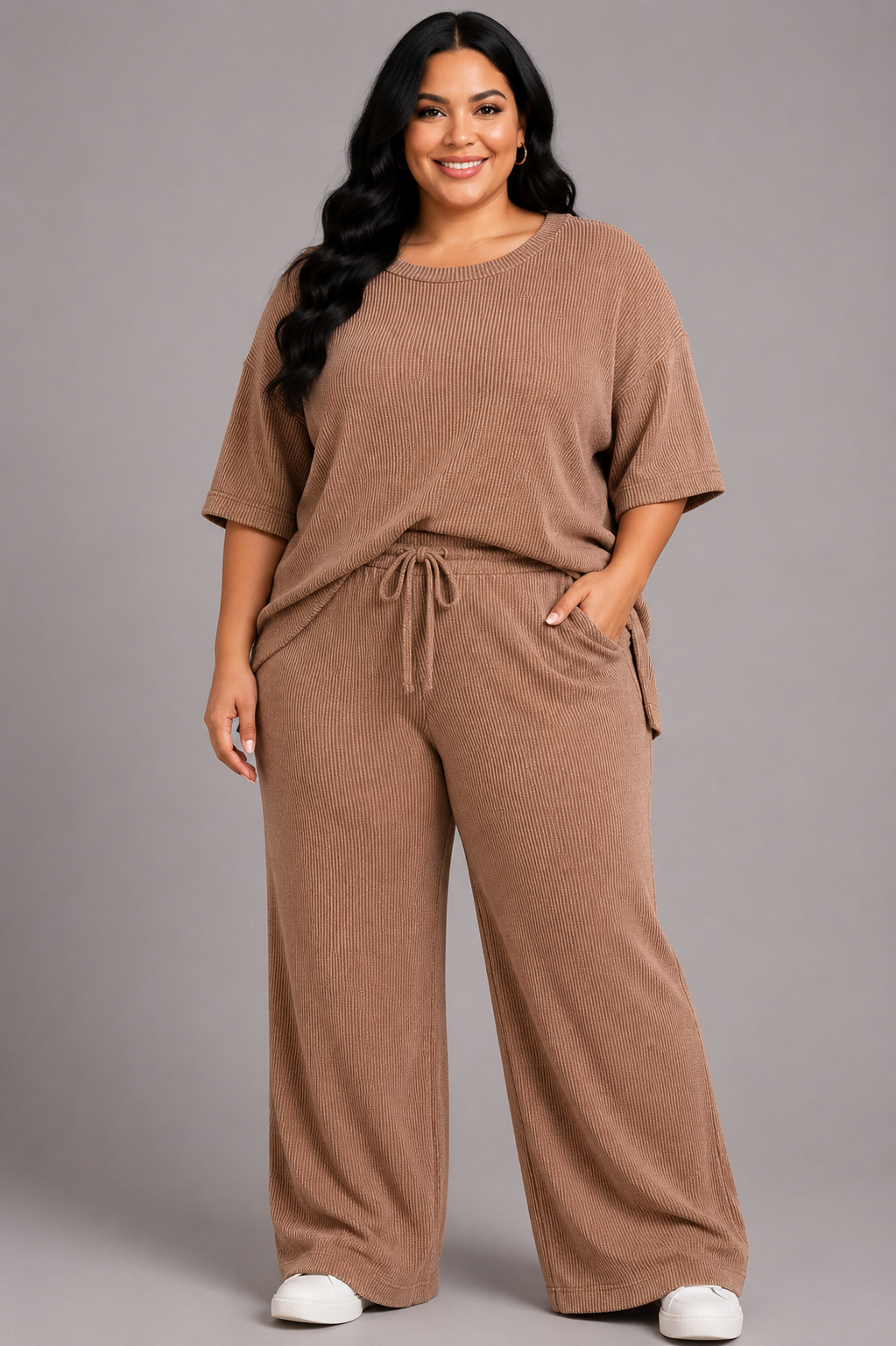 A woman modeling a matching two-piece lounge set in a warm taupe-brown color, featuring a short-sleeve top and wide-leg pants with a drawstring waist, photographed against a solid gray studio background.