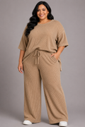 A woman modeling a matching two-piece lounge set in a warm tan color, featuring a short-sleeve crewneck top and wide-leg drawstring pants, set against a solid gray studio background.